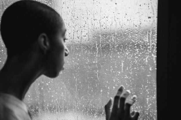 thoughtful androgynous black woman standing near window at rainy day